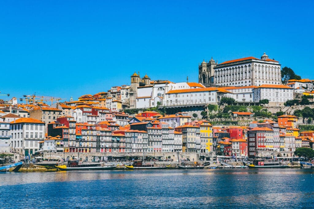 Vista da Cais da Ribeira durante o dia em Porto com rio Douro a frente, alguns barcos dentro do rio e ao fundo várias casinhas.