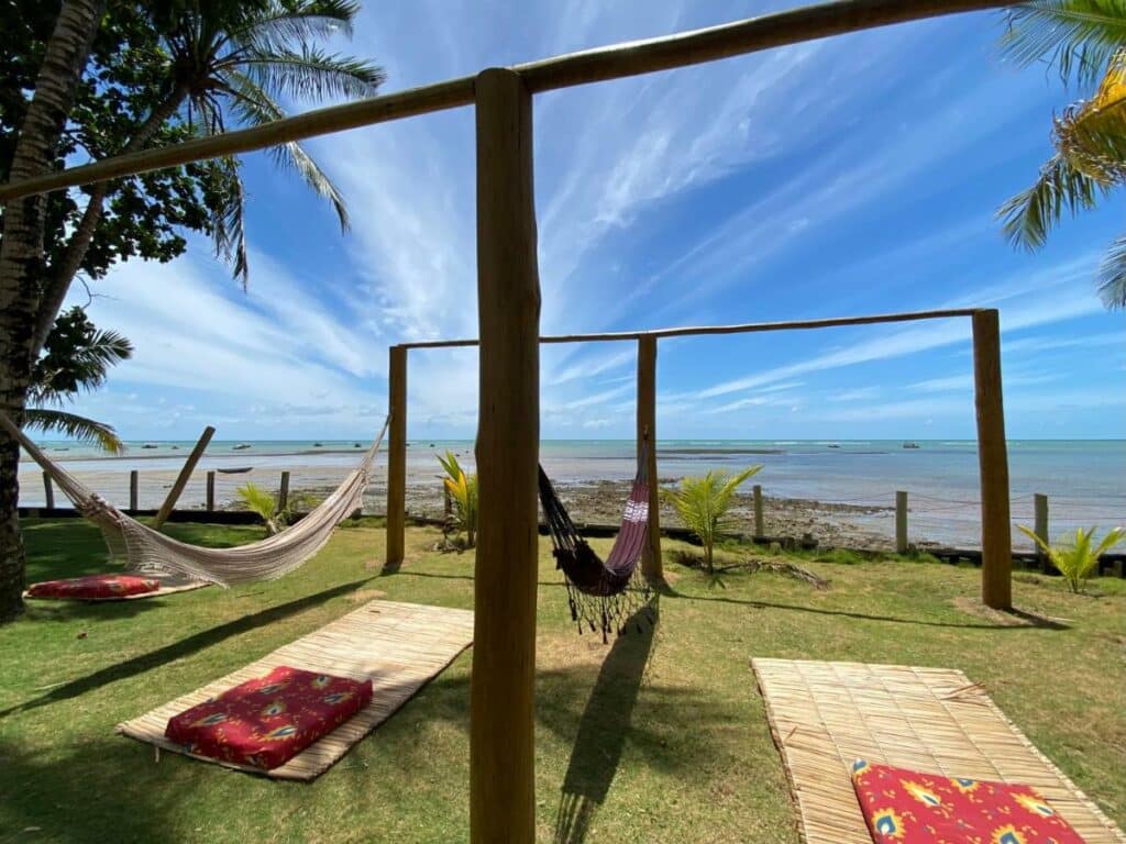 Área externa da Pousada Casarão. Madeiras pendurando duas redes, entre elas esteiras com uma almofada. Atrás a vista para o mar.