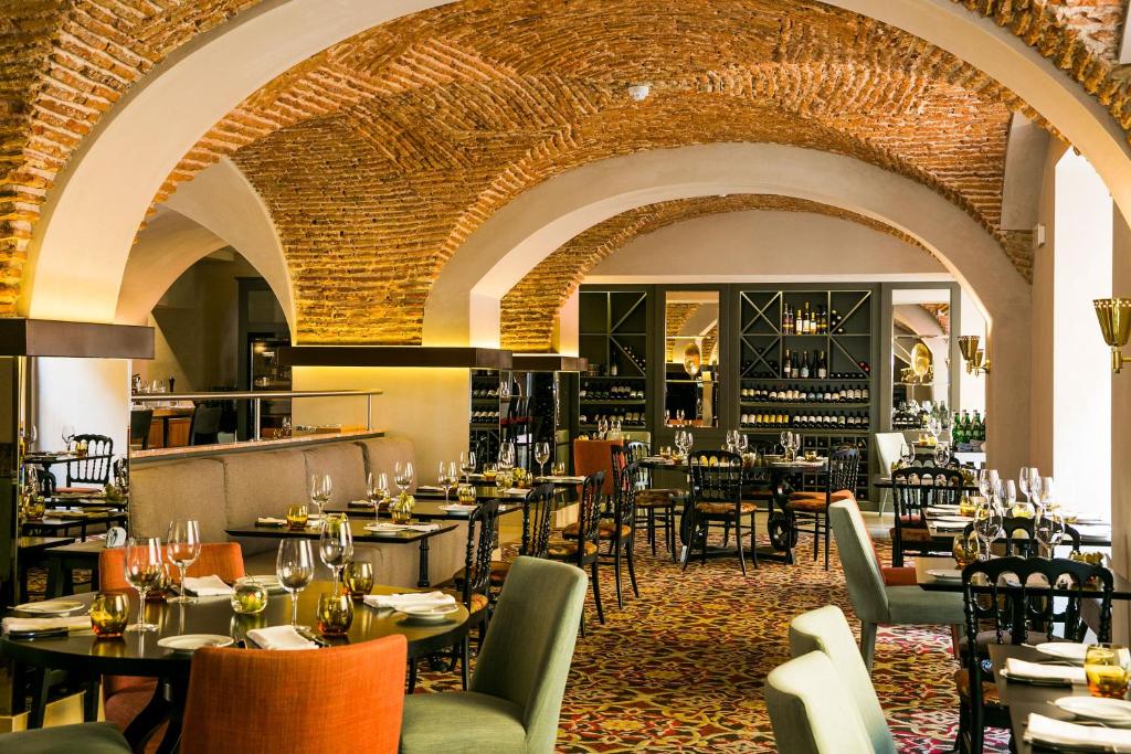 Área de refeições do Pousada de Lisboa - Small Luxury Hotels Of The World com teto de pedra, as mesinhas redondas com cadeiras coloridas, e alguns sofás, ao fundo do local há uma estante com vinhos, para representar hotéis de luxo em Lisboa