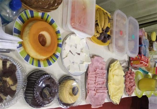 Imagem tirada da mesa de café de manhã da Pousada Líder. Vemos alguns bolos, pudim, frios, sucos, frutas, etc.