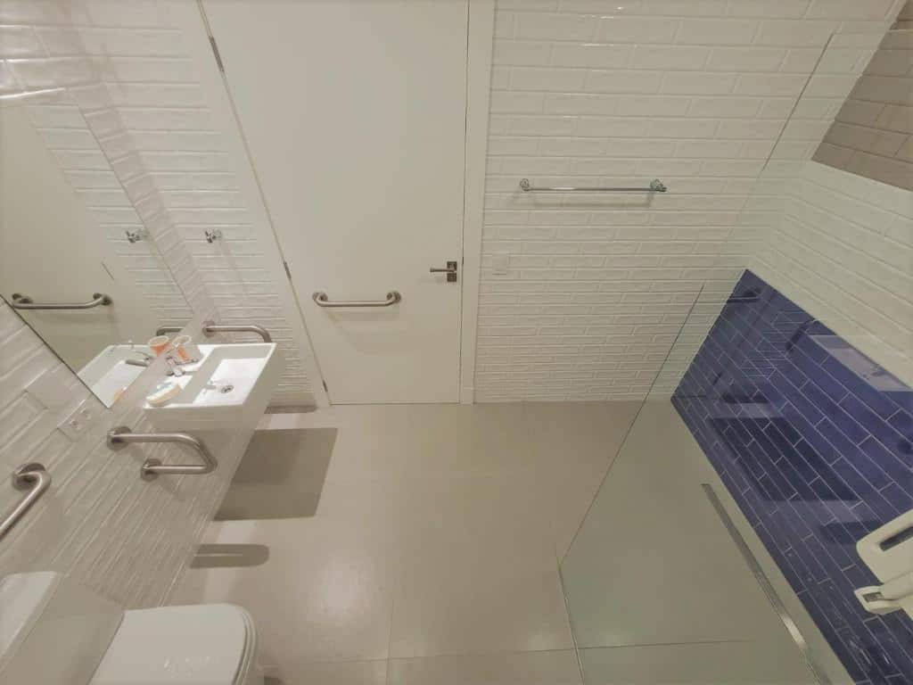 Banheiro do Paranaguá Lodge adaptado para pessoas com baixa mobilidade e deficiência. Do lado esquerdo uma pia com espelho e barras do lado do vaso sanitário. Do lado da pia uma porta com barras. Do lado direito o box com barras.