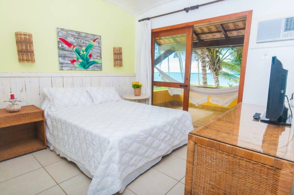 Quarto da Pousada Ponta de Areia. Do lado esquerdo, uma mesa de apoio com um vasinho com flor de cada lado. De frente pra cama uma cômoda e uma televisão, e um ar-condicionado. Atrás, na parede do meio, uma porta de vidro com uma sacada e uma rede e vista para as palmeiras e o mar.