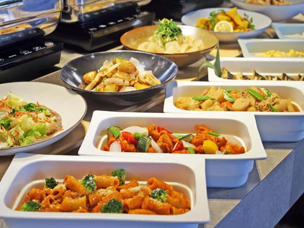 Foto de um buffet servido no Loisir Hotel Shinagawa. É possível ver bandejas e prato com massas, verduras, legumes, etc.