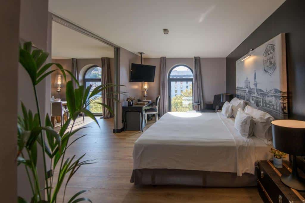Quarto amplo do 138 Liberdade Hotel com uma janela com cortinas, uma cama de casal com travesseiros, uma mesinha de escritório com uma cadeira estofada, com uma televisão em frente da cama, e há uma ante sala ampla, para representar hotéis românticos de Lisboa