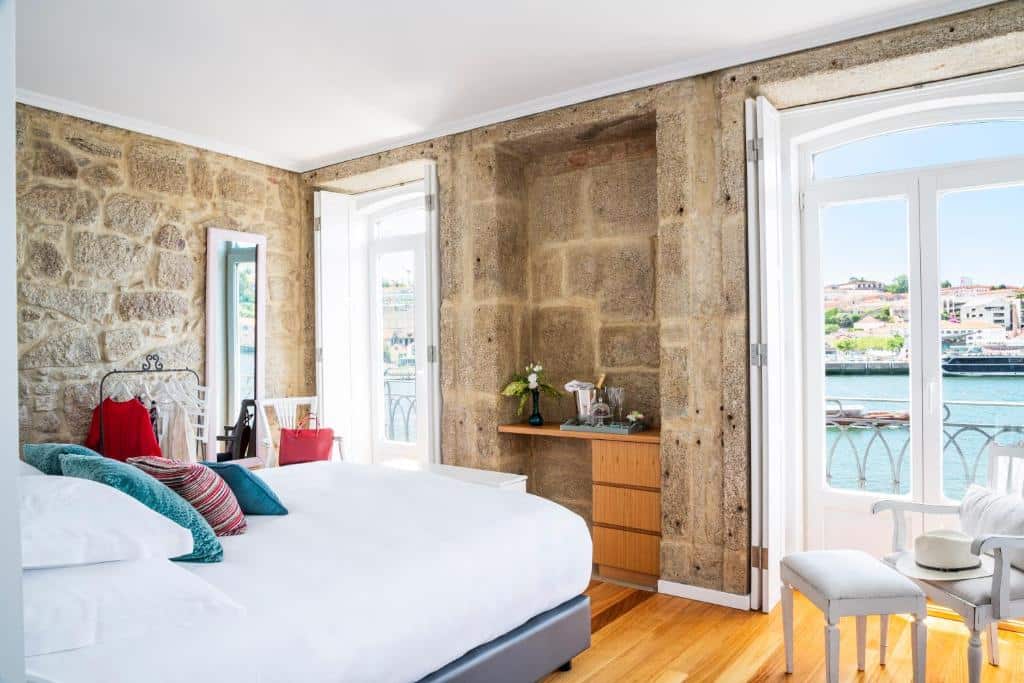 Quarto do 1872 River House com cama de casal do lado esquerdo da imagem, em frente a cama uma cômoda de madeira ao lado de portas que dão acesso a varanda com vista para o rio. Representa hotéis com vista do Douro.