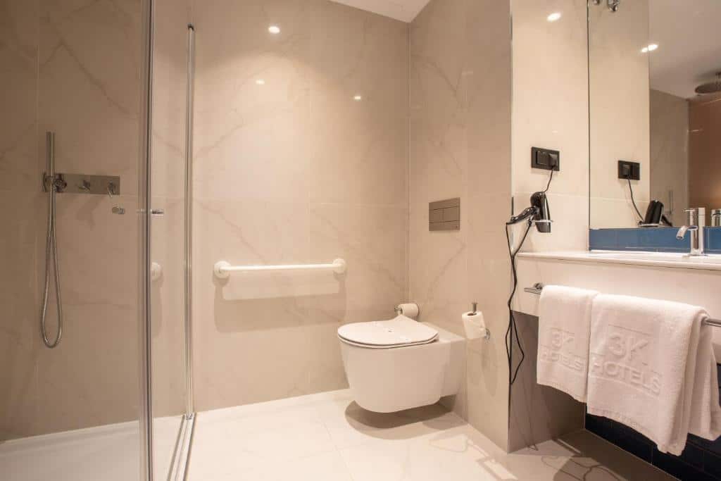 Banheiro com acessibilidade do Hotel 3K Porto Aeroporto com pia d lado direito com barra de apoio, já ao lado da pia vaso sanitária com barras de apoio e em frente ao vaso um box de vidro com chuveiro.