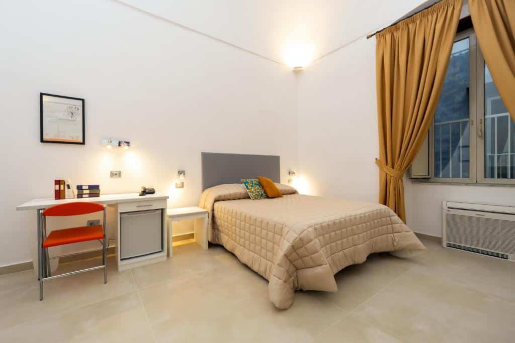Quarto do Il Tesoro Smart Suite & SPA com uma cama de casal, do lado esquerdo uma escrivaninha com livros e objetos em cima, uma cadeira e um frigobar, já no canto direito há um ar-condicionado e uma janela de vidro com cortinas.