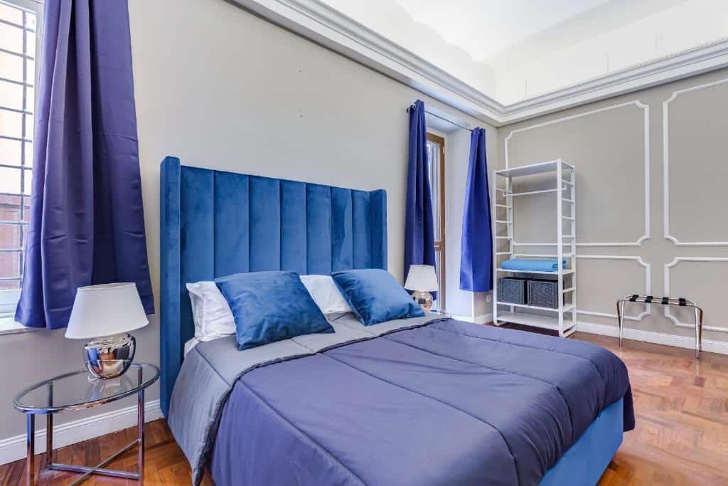 quarto do SPANISH GARDEN, um airbnb em Roma, com cama bem grande, mesinha e luminária de ambos os lados da cabeceira, janela com cortinas azuis combinando com a roupa de cama em um ambiente espaçoso, há uma estante aberta e poltrona branca do outro lado