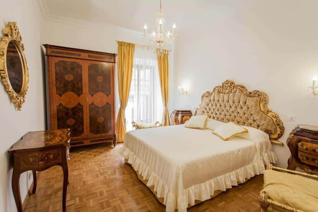 quarto do Suite Sarandrea, aluguel de temporada em Roma, com cama enorme com decorações clássicas, mesinha e luminárias de ambos os lados, além de um lustre acima, guarda-roupa e móveis de madeira, e janela grande com cortinas