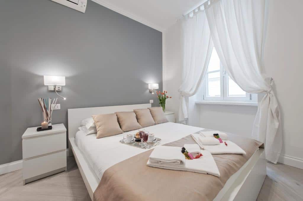 quarto do Trevi Apartment, um aluguel de temporada em Roma, com cama grande e detalhes em rosê e cinza, além de branco, há uma janela com cortinas leves, luminária e mesinha de ambos os lados da cabeceira e ar-condicionado