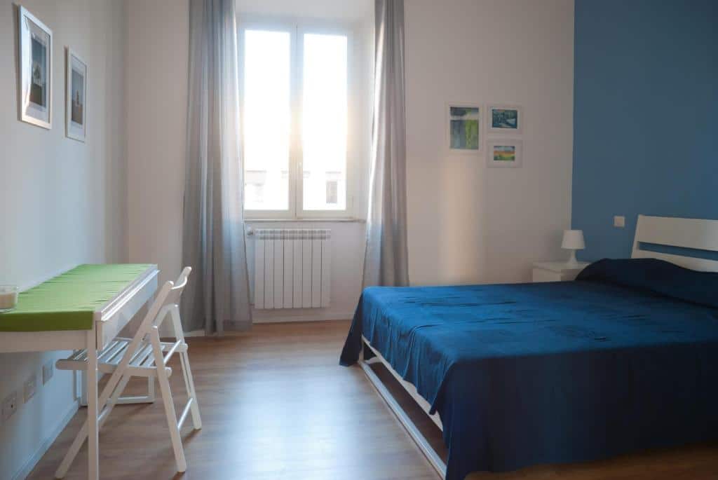 quarto do Welcome Friends, um airbnb em Roma, com cama de casal, mesinha de cabeceira e luminária de ambos os lados, mesa com cadeira à frente e, do outro lado, uma janela com cortinas e aquecedor