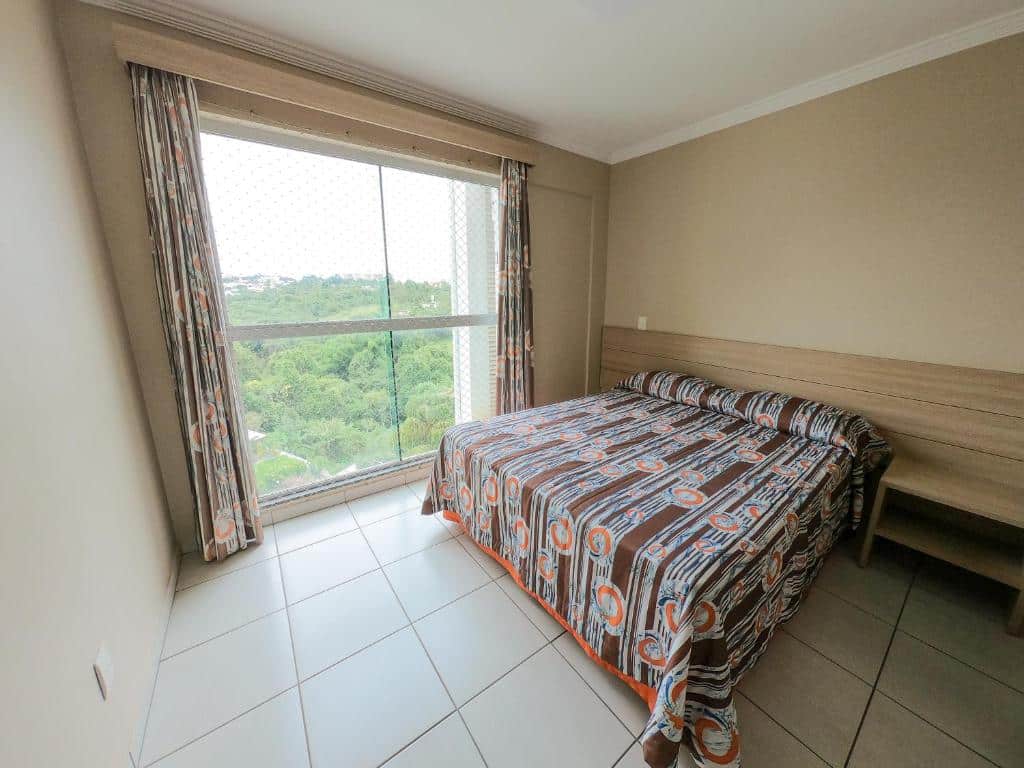 Quarto do hotel Águas do Paranoá com uma cama de casal, com uma mesinha de madeira ao lado da cama e uma janela grande de vidro com vista para as árvores.