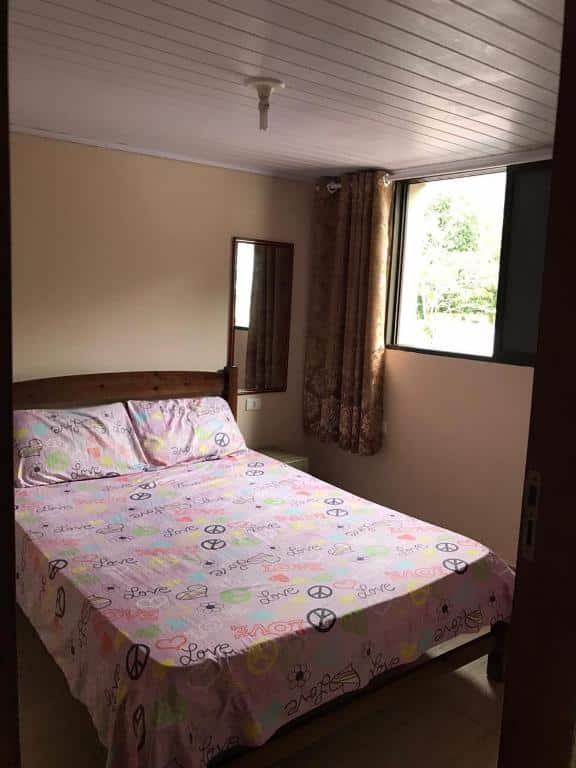 Quarto pequeno com paredes em tom de rosa claro, cama de casal centralizada, pequeno espelho no lado direito da cama, janela e cortina também no lado direito. Imagem para ilustrar o post pousadas em Mongaguá.