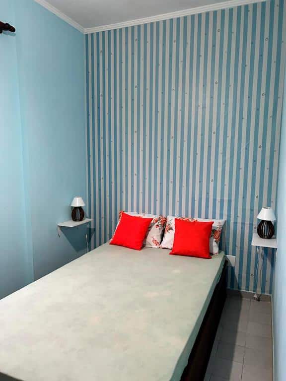 Pequeno quarto com cama de casal centralizada e pequenas prateleiras com luminárias em cada lado da cama. Imagem para ilustrar o post pousadas em Mongaguá.