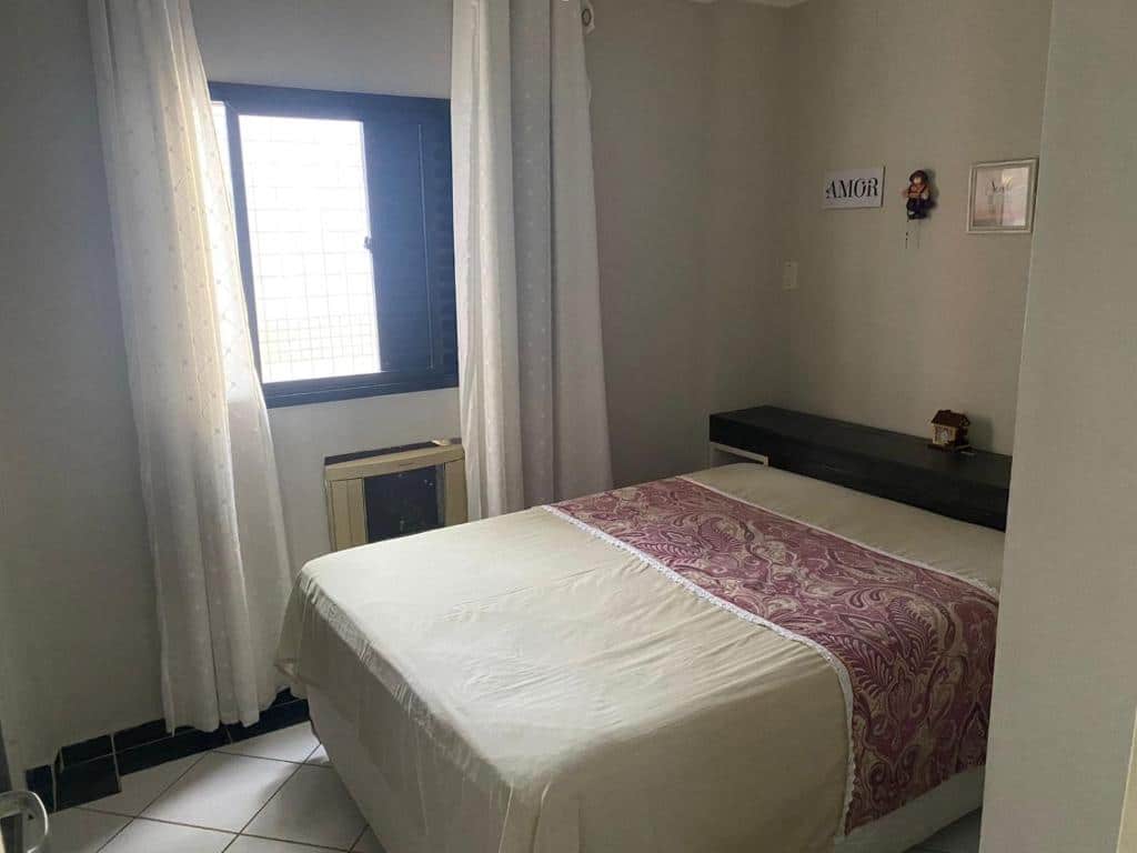 Quarto com janela, cortina e ar condicionado na parede esquerda e cama de casal centralizada. Imagem para ilustrar o post pousadas em Mongaguá.