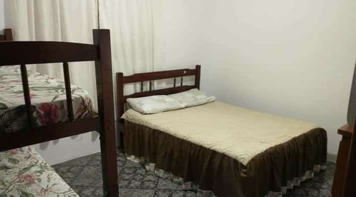 Quarto com beliche no lado esquerdo, cortina branca centralizada e cama de casal no lado direito. Imagem para ilustrar o post pousadas em Mongaguá.