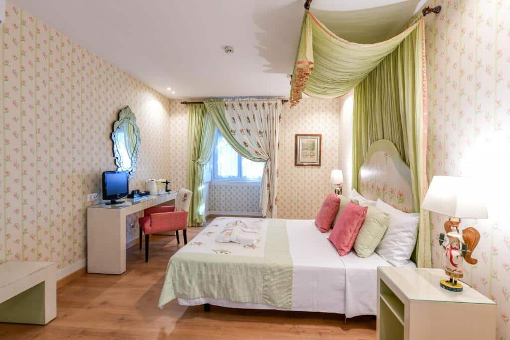 Quarto do Casa de Sao Mamede Hotel com uma cama de casal com um bangalô no alto com um tecido verde claro, há um janela com cortinas no mesmo tom, de frente para a cama há uma bancada com uma televisão e um espelho, além de uma poltrona, o chão do quarto imita madeira, para representar hotéis baratos em Lisboa