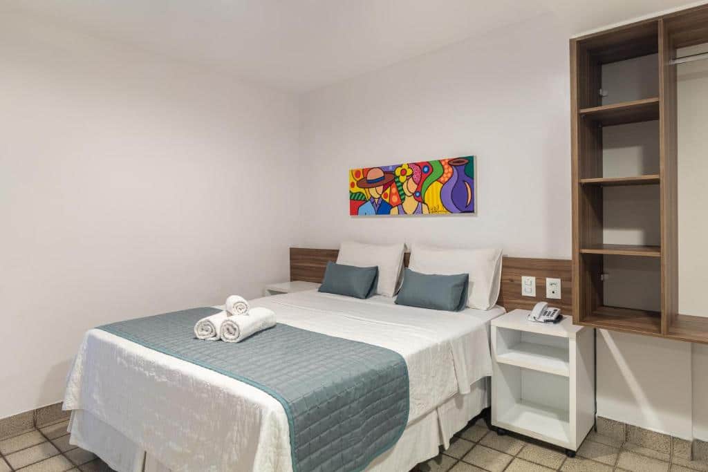 Quarto do Citi Hotel Express Caruaru. A cama está encostada no lado direito com um pequeno móvel no lado direito e no lado esquerdo da cama e mais a direita é possível visualizar prateleiras de um armário. Imagem para ilustrar o post hotéis em Caruaru.