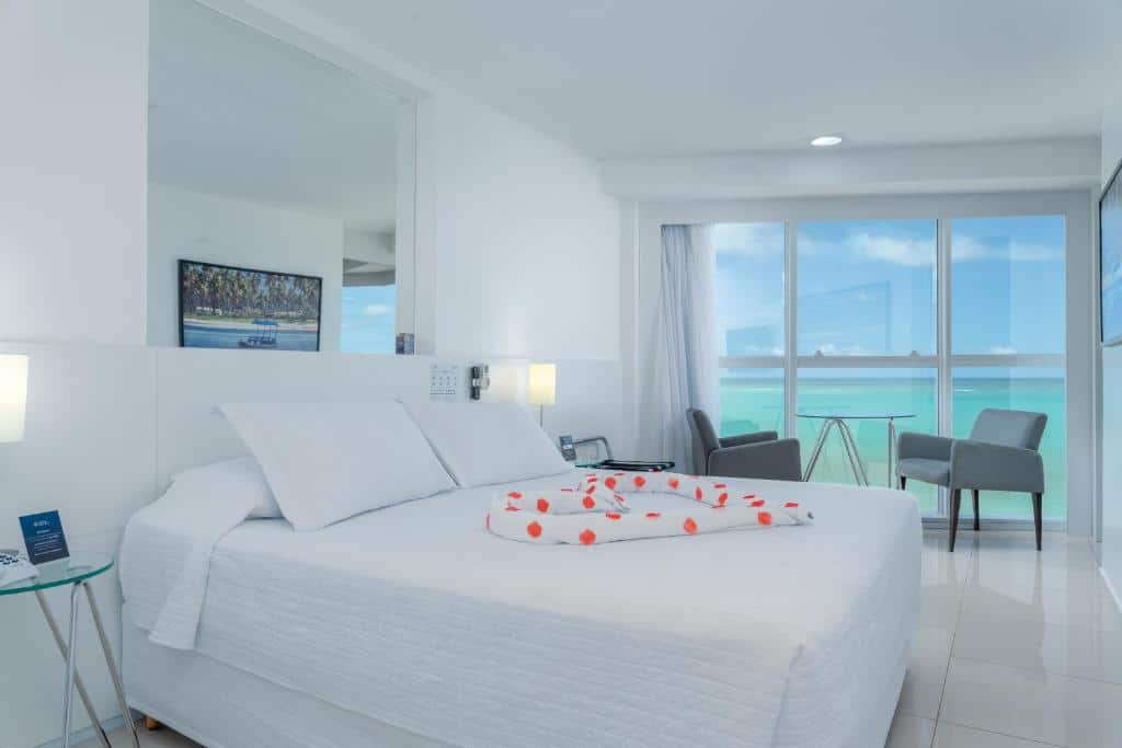 Quarto do Hotel Brisa Praia, de 40 m², em Maceió, com uma cama de casal com um coração no meio feito de toalhas enroladas e pétalas vermelhas, uma mesa redonda com duas poltronas e, ao lado, uma janela panorâmica com vista do mar