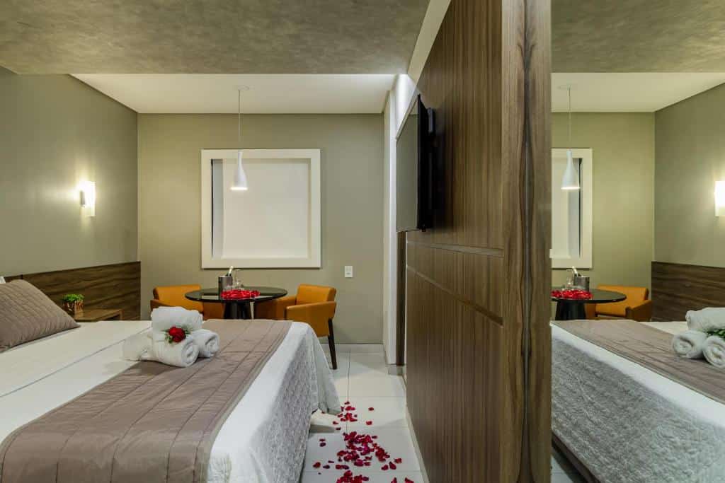 Quarto do Citi Hotel Premium Caruaru. A cama de casal está encostada no lado esquerdo com toalhas em cima. Ao fundo há uma mesa redonda com duas cadeiras. No lado direito a TV está presa na parede e tem pétalas espalhadas pelo chão. Imagem para ilustrar o post hotéis em Caruaru.