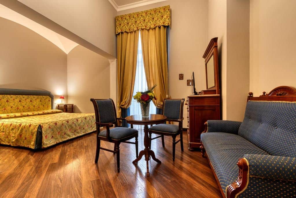 Quarto do Decumani Hotel De Charme com uma cama de casal na parte esquerda ao fundo, em frente uma mesinha redonda com um vaso de flor e duas cadeiras, no lado direito um sofá. Há também um móvel de madeira e uma janela de vidro com cortinas.