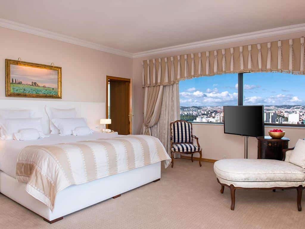 Quarto do Dom Pedro Lisboa com uma cama de casal, uma janela ampla com cortinas, há uma televisão com uma poltrona ao lado, o chão é de carpete bege
