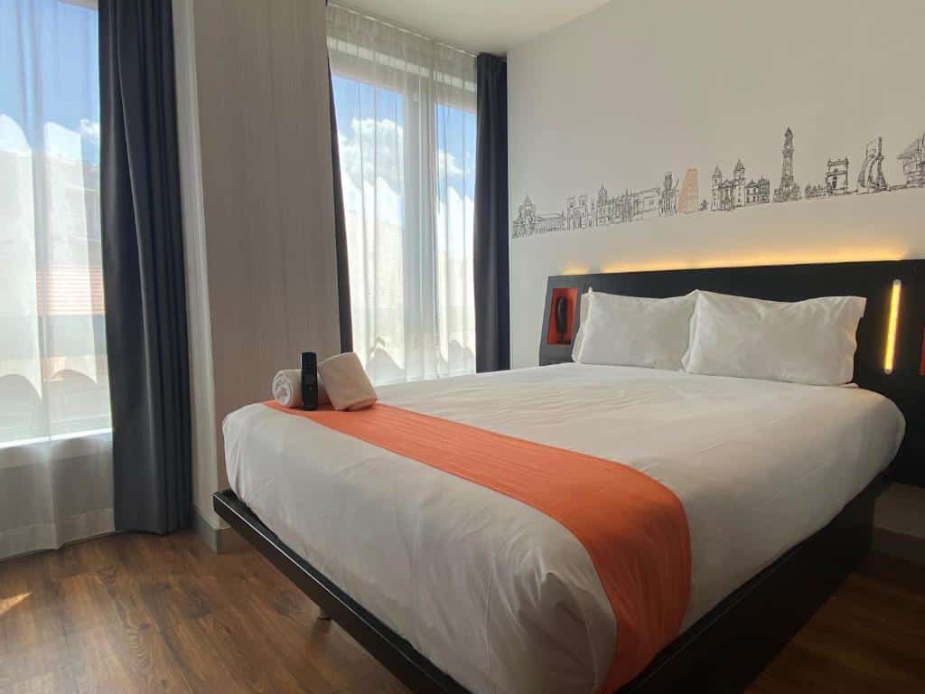 Quarto do easyHotel Lisbon com uma cama de casal, do lado esquerdo, há duas janelas com cortinas brancas e o chão do ambiente imita madeira