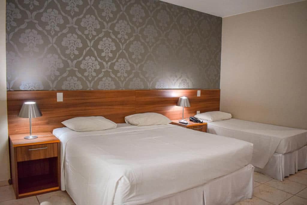 Quarto do Ecologic Ville Resort com cama de casal do lado esquerdo da imagem, uma mesa com luminária ao centro e uma cama de solteiro do lado direito.