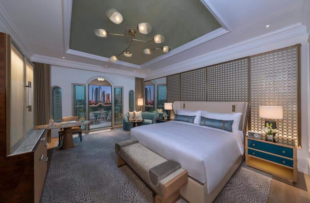 Quarto do Emirates Palace Mandarin Oriental, Abu Dhabi. Uma cama de casal no lado direito, com uma cômoda com abajur de cadal lado e um sofá na frente. De frente para a cama um armário. No fundo do quarto,no lado esquerdo uma mesa de trabalho e no lado direito um sofá. Atrás, a sacada do quarto.