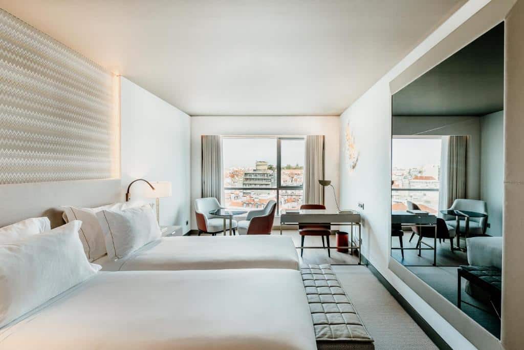 Quarto do EPIC SANA Marquês Hotel com duas camas de solteiro, uma varanda com cortinas com vista para a cidade, perto da janela há uma pequena mesinha com suas poltronas e uma mesa de escritório, além de um amplo espelho de frente para as camas