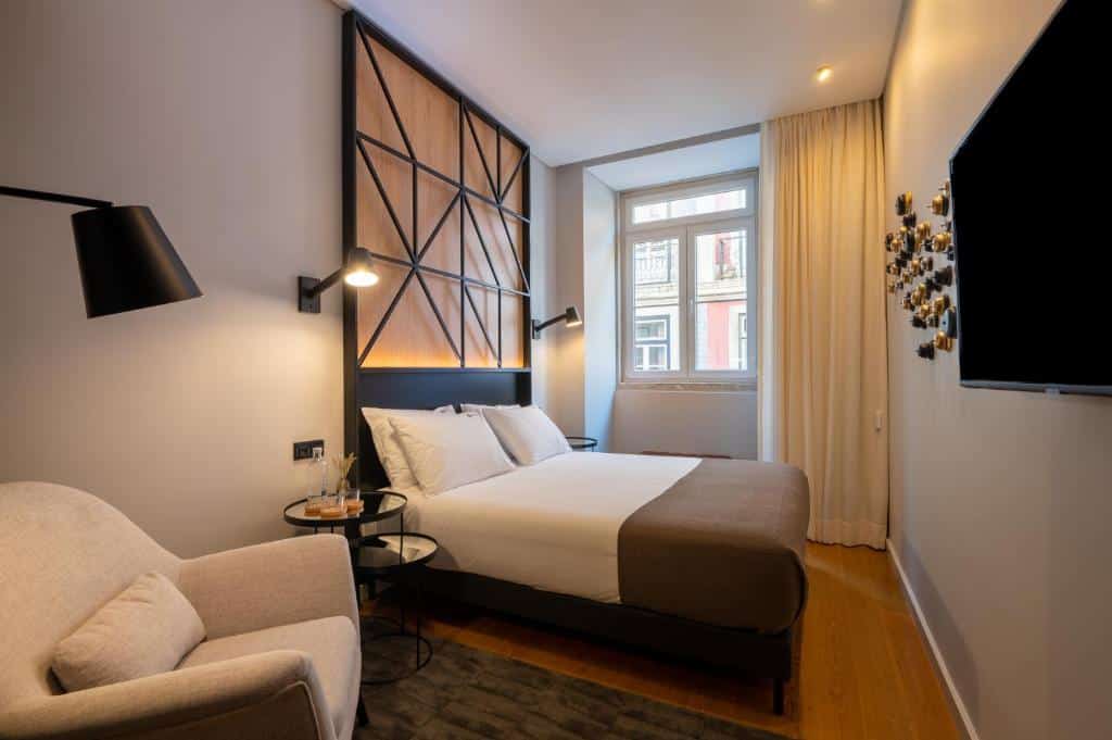 Quarto do Ferraria XVI FLH Hotels Lisboa  com uma cama de casal com travesseiros, ao lado direito da cama há uma janela com cortinas, em frente da cama há uma televisão presa na parede, e do lado esquerdo há uma poltrona e uma luminária de chão, para representar hotéis no Chiado em Lisboa