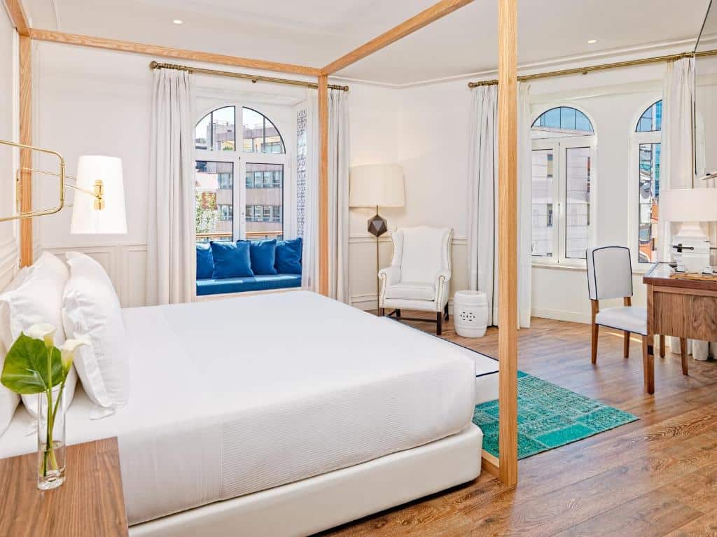 Quarto chique do H10 Duque de Loule com uma cama de casal ampla, duas janelas, sendo que numa delas há um pequeno sofá com almofadas, há também uma poltrona branca com um abajur de chão, para representar hotéis bem localizados em Lisboa