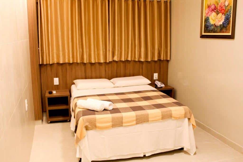 Quarto do Vila Rica Hotel Caruaru. A cama está centralizada, há um pequeno móvel de madeira no lado direito e no lado esquerdo da cama. Uma cortina fechada está localizada em cima da cama. Imagem para ilustrar o post hotéis em Caruaru.