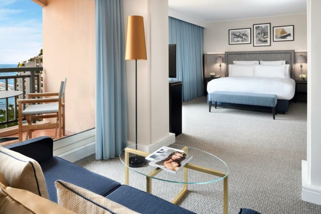 Quarto do Hôtel Columbus Monte Carlo. Um sofá na frente com uma mesa no centro, atrás o quarto, com uma cama de casal e de frente pra cama uma televisão. No canto esquerdo, do lado do sofá, a varanda do quarto.