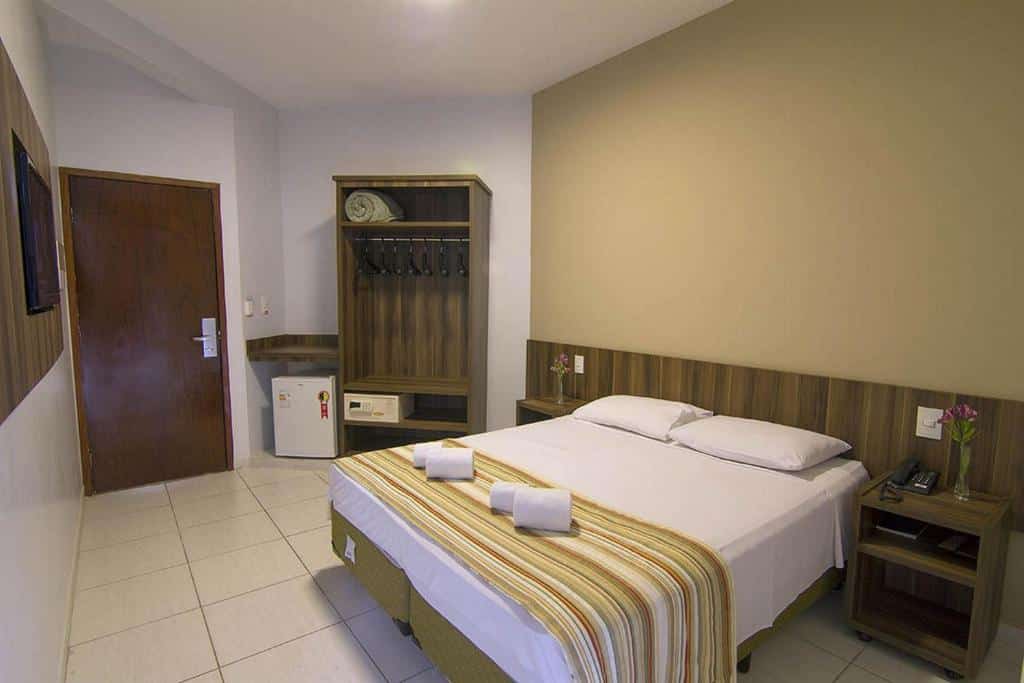 Quarto do Hotel Morada das Águas com uma cama de casal, móveis em madeira, guarda-roupa com cofre e cabides, um frigobar ao lado e uma tv na parede.