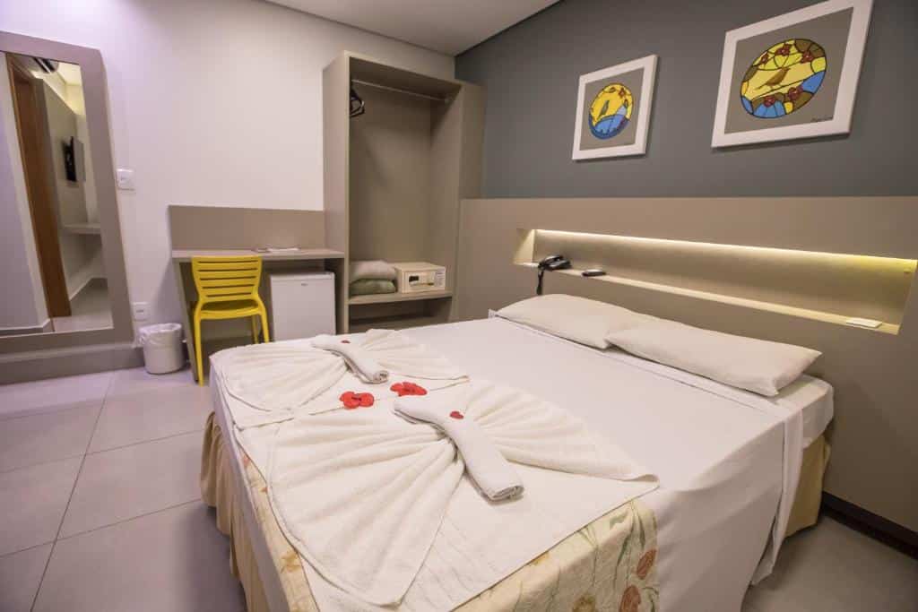 Quarto do Hotel Morada do Sol com uma cama de casal, dois quadros na parede, um guarda-roupa aberto com espaço para as roupas e um cofre, ao lado uma escrivaninha com um frigobar e uma cadeira.