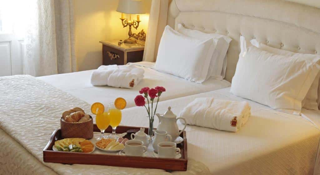 Cama de um dos quartos do Hotel Real d Obidos com cobertas, travesseiros e roupões brancos, sob a mesma há uma bandeja com itens de café da manhã, como pães e frutas