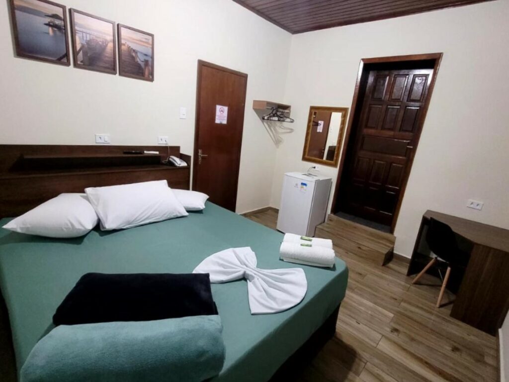 Quarto do Hotel Serra do Mar. Uma cama de casal no lado esquerdo, no lado direito um frigobar, um espelho, a porta do quarto e uma mesa de trabalho.