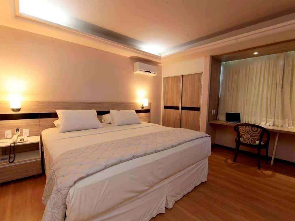 Quarto do Hotel Serrano. Uma cama de casal no lado esquerdo, em cada lado da cama uma prateleira com luminária. No lado direito da cama um ar-condicionado e a porta do guarda-roupa. No lado direito do quarto uma mesa de trabalho de frente pra janela do quarto.