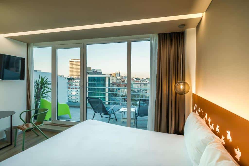 Quarto do Hotel Ibis Styles Lisboa Marquês de Pombal com varanda com cadeiras e uma vista da cidade, no quarto, há uma cama de casal, uma televisão e uma pequena mesinha com uma cadeira