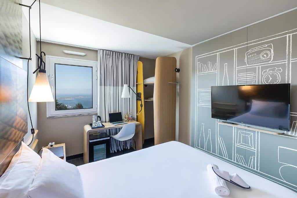Quarto do Hotel ibis Porto Gaia com cama de casal do lado esquerdo da imagem, em frente a cama uma TV presa na parede e do lado esquerdo da cama uma mesa de trabalho. Representa hotéis ibis no Porto.