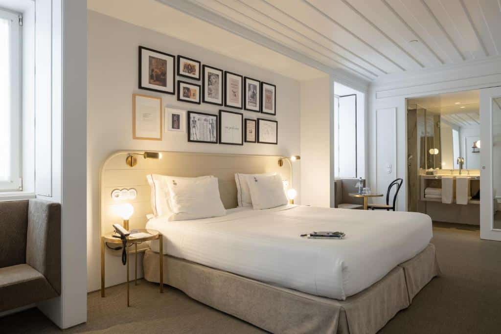 Quarto do Lisboa Pessoa Hotel com uma cama de casal, na parede atrás da cama há diversos quadros, nas laterais da mesma há mesinhas com luminárias, há duas janelas do cômodo, para representar hotéis no Chiado em Lisboa