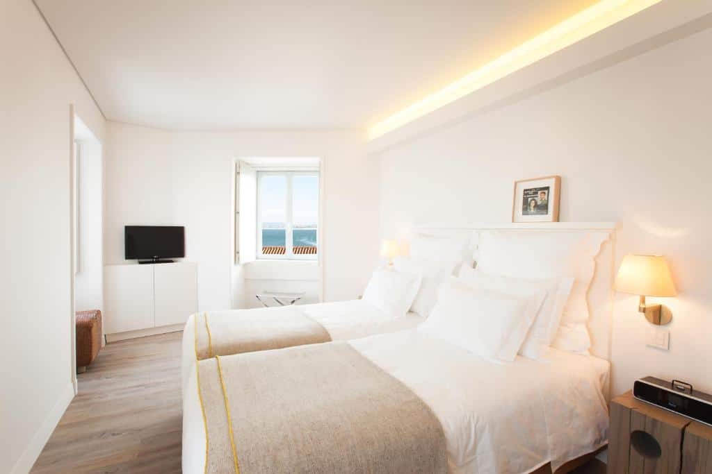 Quarto do Memmo Alfama - Design Hotels com duas camas de solteiro, uma pequena janela com vista para o mar, ao lado da janela há um pequeno móvel branco com uma televisão em cima, nas laterais da cama há luminárias presas na parede