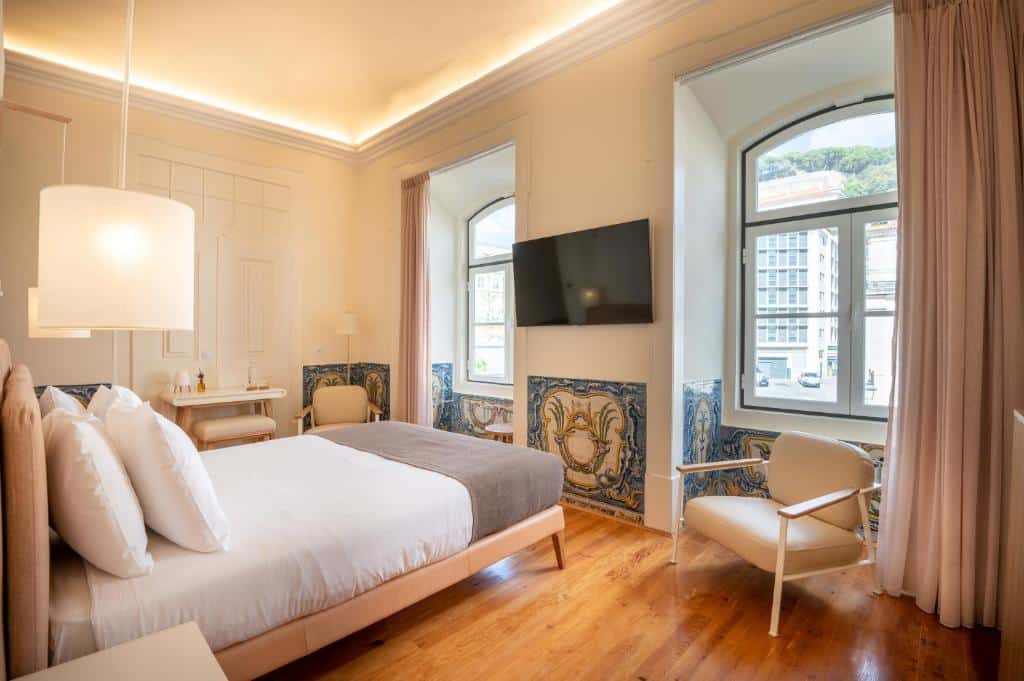 Quarto do Memoria Lisboa FLH Hotels com uma cama de casal de frente para uma parede com duas janelas e, entre elas, há uma televisão presa, do lado esquerdo do quarto há uma pequena bancada e uma cadeira estofada, o chão imita madeira e algumas paredes tem azulejos coloridos