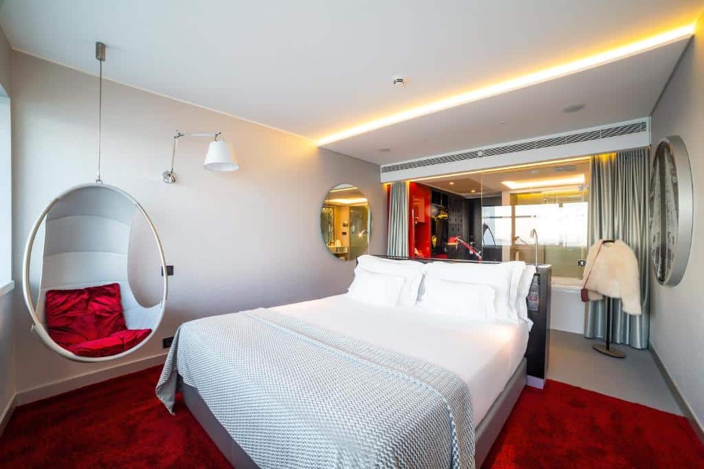 Quarto do MYRIAD by SANA Hotels com uma cama de casal, uma cadeira pendurada no teto, dois espelhos, um tapete vermelho e um vidro separando o banheiro do quarto
