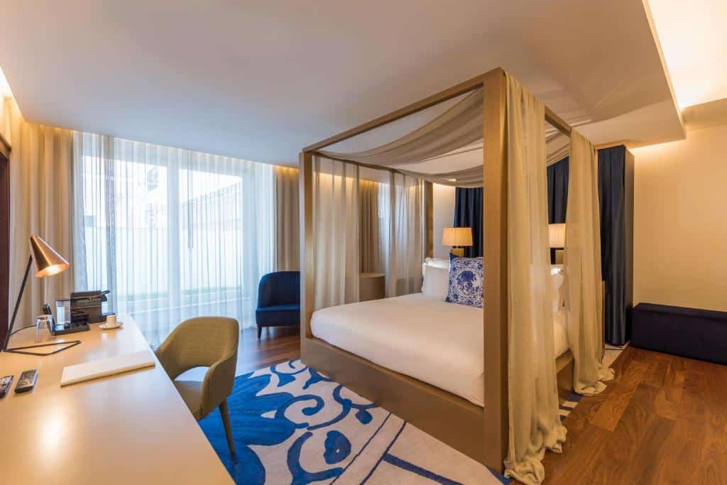 Suíte espaçosa do NAU Palacio do Governador com uma cama de casal com um bangalô com tecidos, há uma janela ampla com cortinas, há também uma mesa de escritório com uma cadeira e um tapete branco e azul, para representar hotéis bem localizados em Lisboa