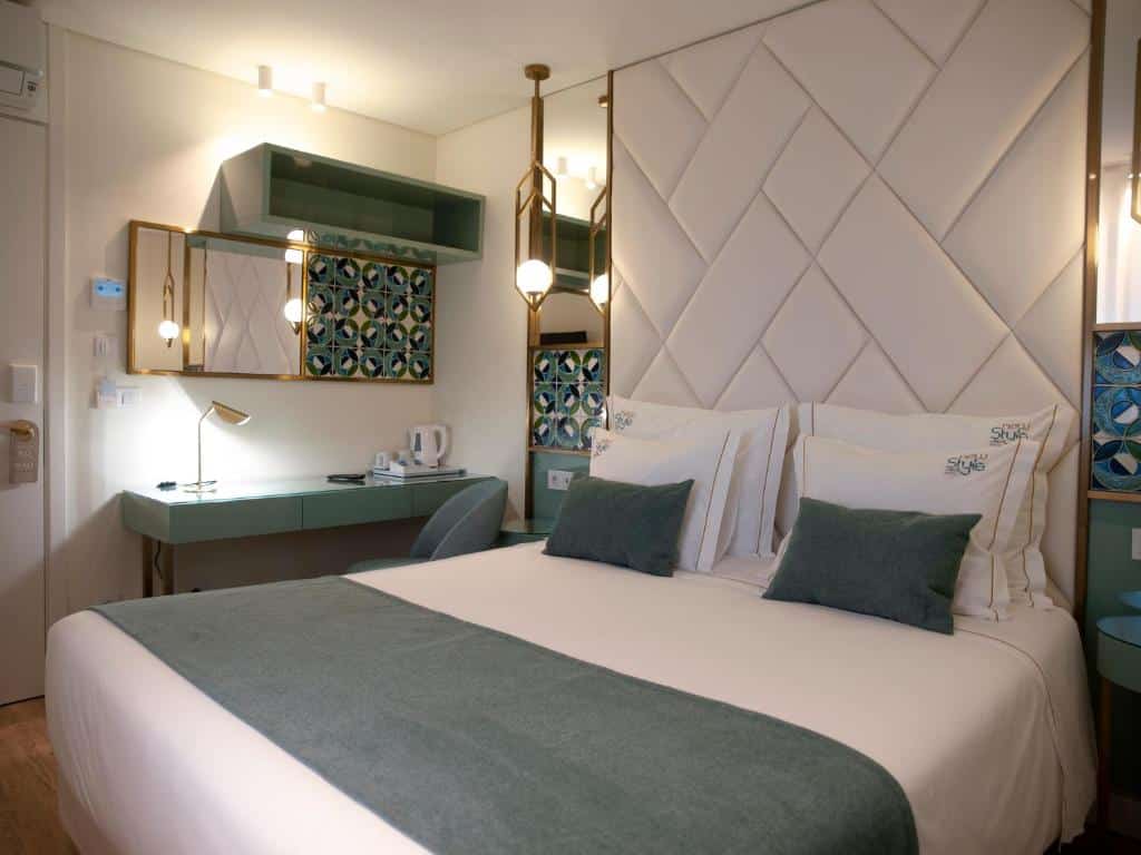 Quarto do New Style Lisbon Hotel com uma cama de casal, a cabeceira é estofada e conta com pequenos espelhos nas laterais, do lado esquerdo da cama é possível ver uma mesa de escritório com uma cadeira