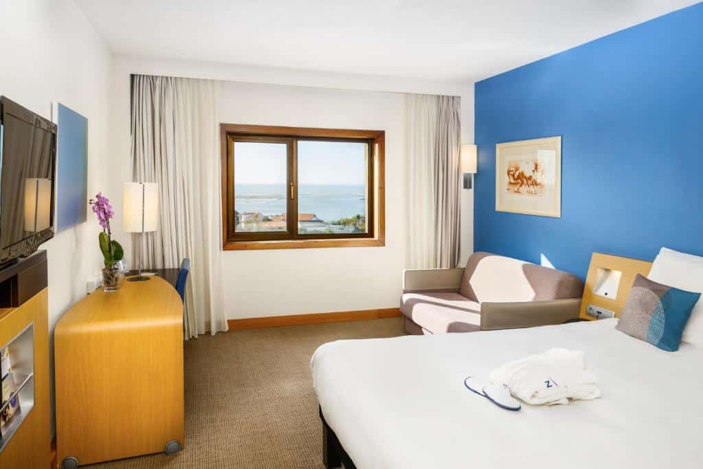 Quarto do Novotel Porto Gaia com cama do lado direito da imagem a frente, do lado esquerdo da cama um sofá de dois lugares e em frente a cama uma TV em cima de uma cômoda. Representa hotéis ibis no Porto.