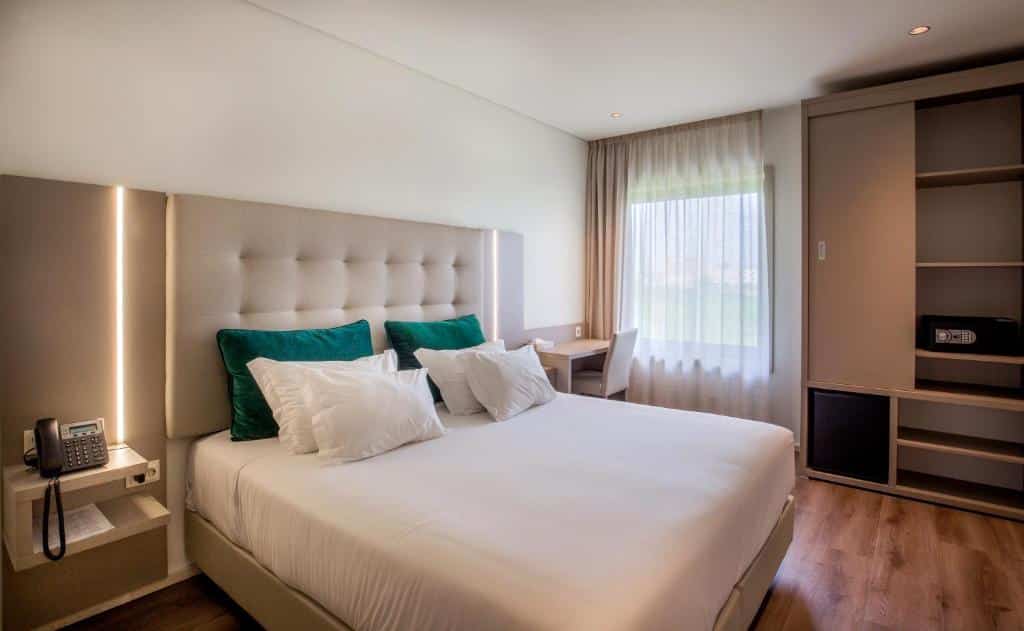Quarto do Oporto Airport & Business Hotel com cama de casal do lado esquerdo do quarto, do lado esquerdo da cama um telefone numa cômoda e do lado direito da cama uma mesa de trabalho com cadeira. Representa hotéis perto do aeroporto do Porto.