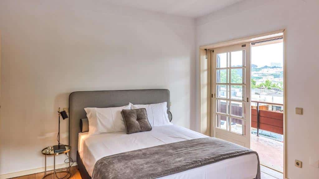 Quarto do Porto River House com cama de casal do lado esquerdo da imagem, do lado esquerdo da cama uma mesinha com luminária e do lado direito uma porta de vidro que dá acesso a varanda.